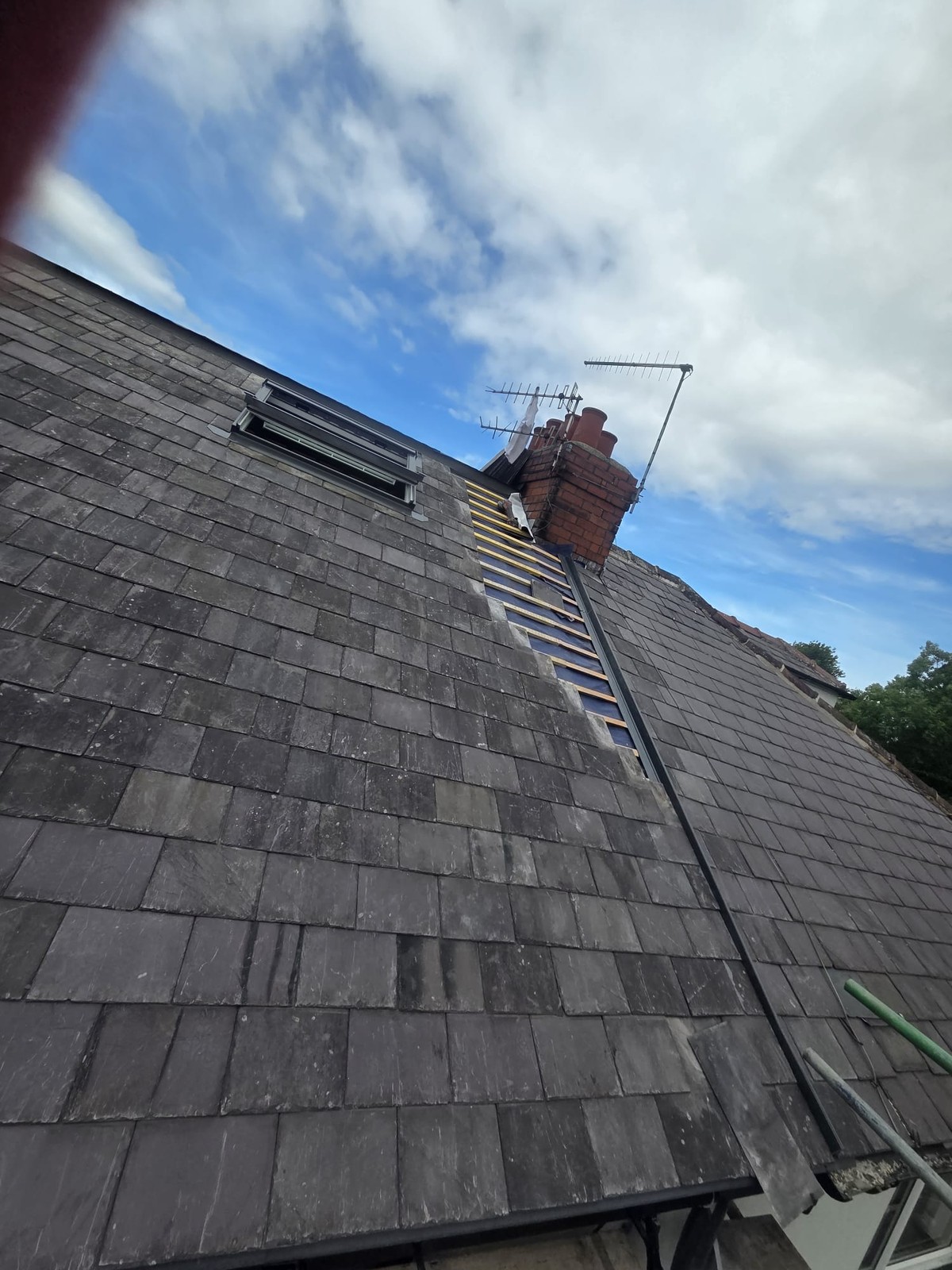 Access ladder resting on a slate roof below a brick chimney, with a roof window nearby